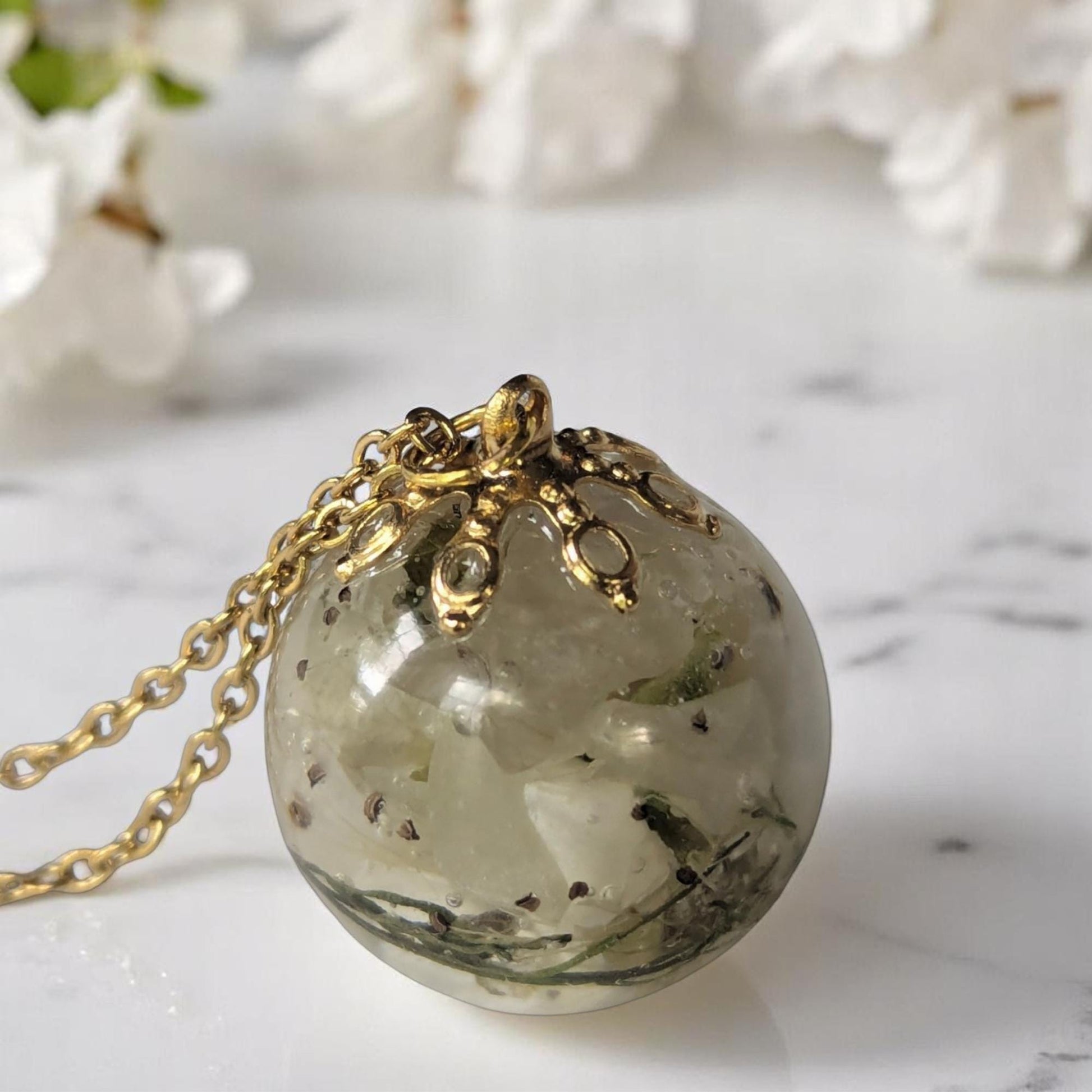 A gold plated necklace chain with a spherical pendant containing dried Bradford Pear flower blooms, set against a marble background with white flowers.
