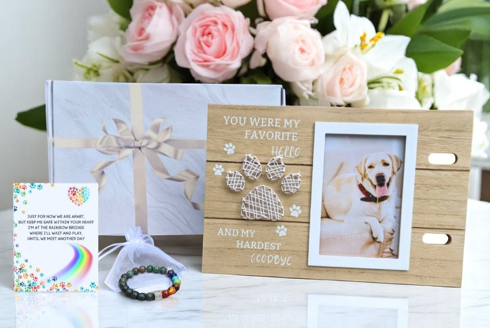 Wooden photo frame with a dog picture, gift box, card, and bracelet on a table with flowers in the background.