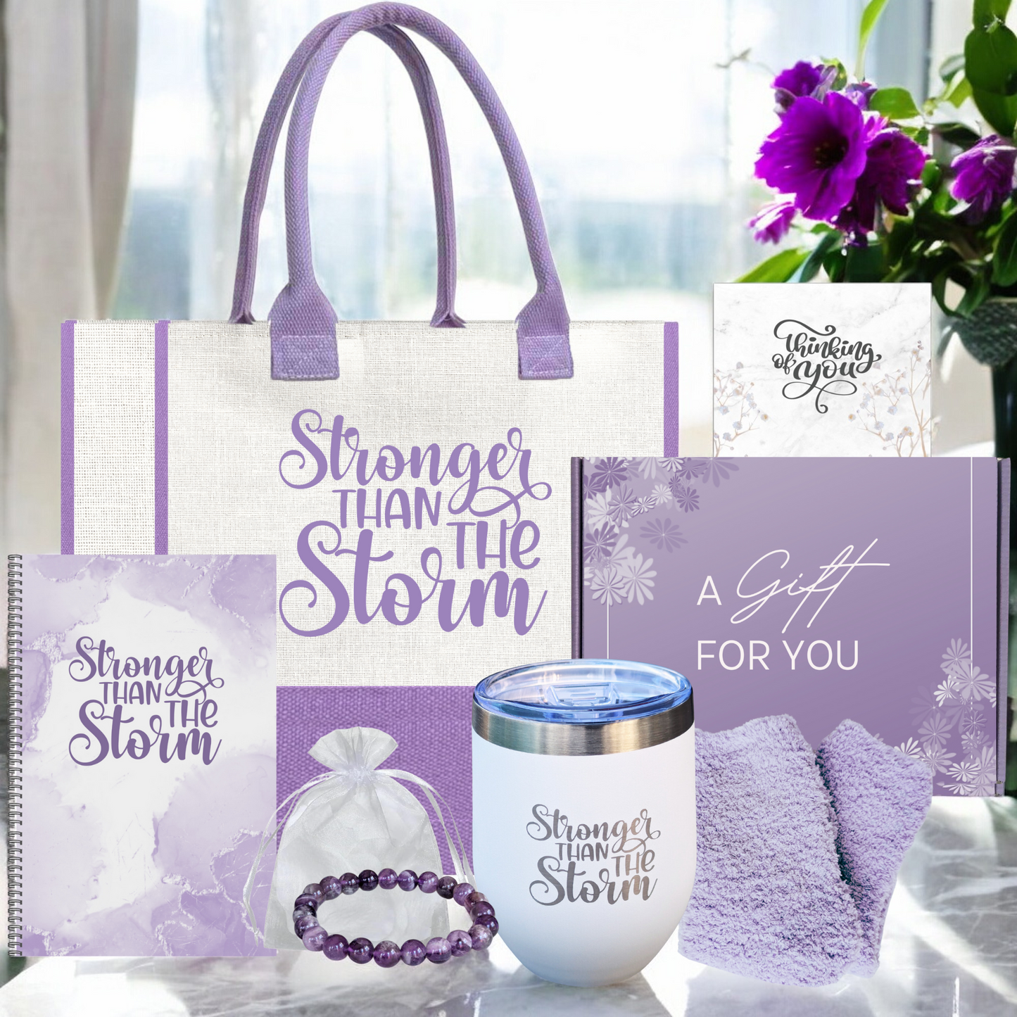 A gift tote bag with 'Stronger Than The Storm' printed, alongside a white insulated tumbler, a purple stone bracelet, cozy socks, a spiral notebook, and a thinking of you card.